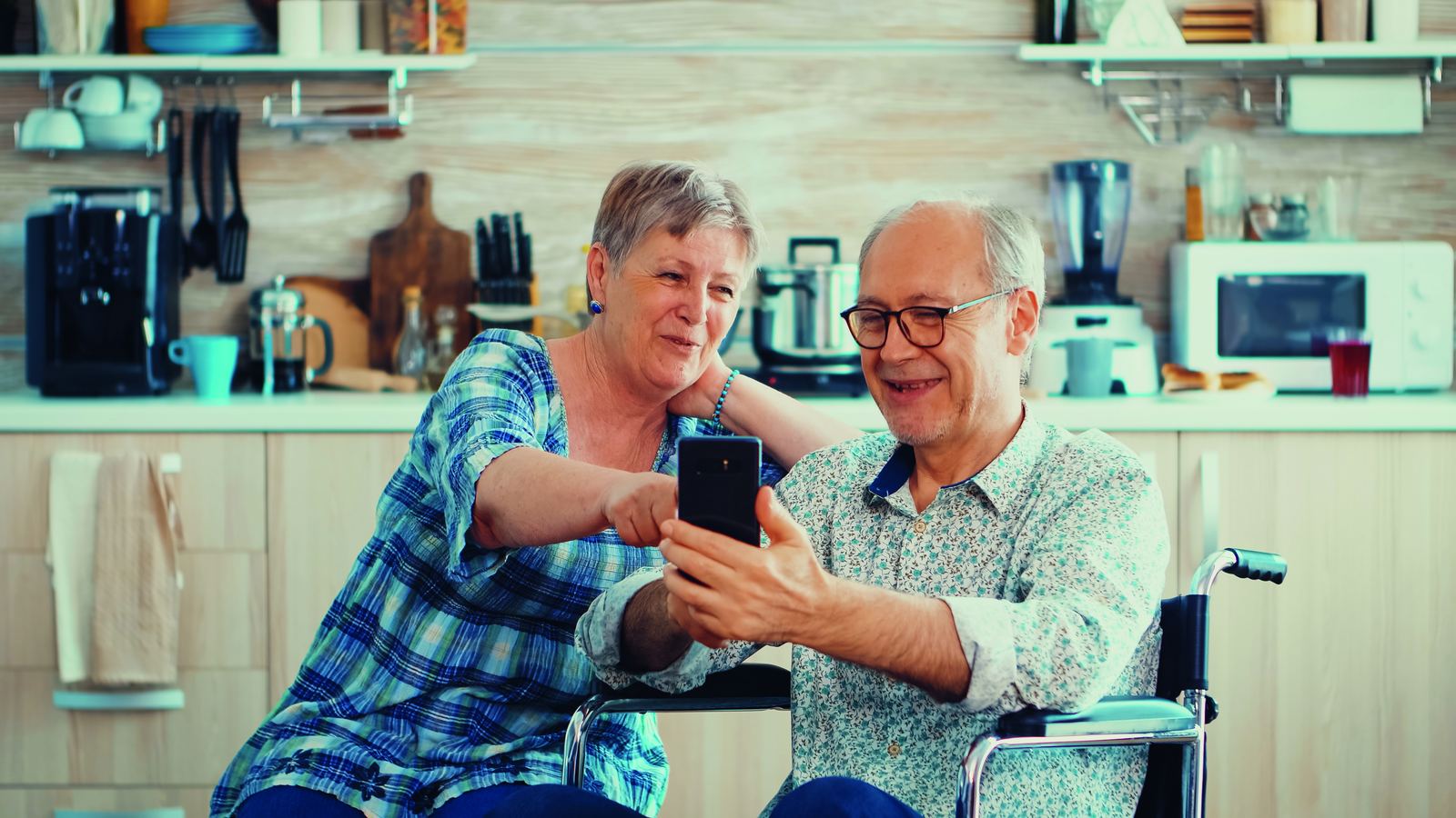 Seniorin und ihr Ehemann im Rollstuhl nutzen gemeinsam ein Smartphone in einer Küche.